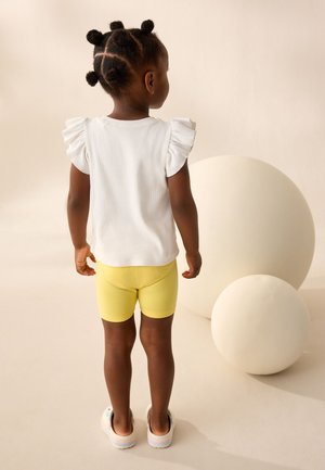 Ungt barn med flettet hår står med fronten væk, iført en hvid bluse med flæser på ærmerne, gule shorts og hvide sko mod en neutral baggrund.