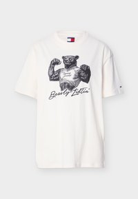 Λευκό μπλουζάκι με έναν μυώδη αρκούδο που λυγίζει τα μπράτσα του, φορόντας φανελάκι με την ένδειξη «Tommy Jeans» και το κείμενο «Bearly Liftin'» από κάτω.