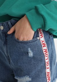 Shorts de mezclilla con efecto desgastado, que presentan un diseño rasgado y una etiqueta tejida roja y blanca que dice "SIGUE MI CAMINO" a lo largo de la costura lateral.