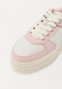 Baskets avec une tige en toile blanche et rose pâle, dotées d'une semelle en caoutchouc, d'un bout rond et de lacets blancs sur une surface texturée.