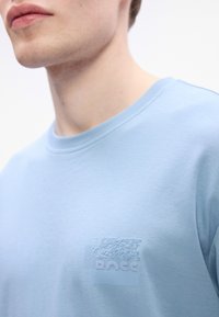 Jauns vīrietis, ģērbies gaiši zilā apģērbtajā kreklā ar izcilu logo kreisajā krūtīs, redzams no pleciem līdz mutei, uz baltā fona.