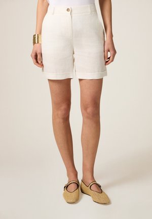 Shorts en lin de couleur crème clair, avec un ourlet roulé, des poches latérales et une fermeture à bouton. Associés à des chaussures à enfiler en tressé.