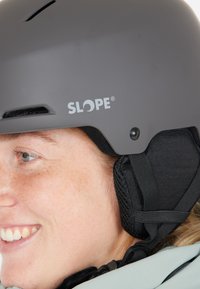 Ung person iført en grå Slope hjelm med sorte polstrede ørebeskyttere, smilende og kigger til siden.