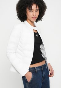 Fehér párnázott dzseki magas gallérral, elülső cipzárral és steppelt textúrával. Alatta fekete trikó, kék farmerrel párosítva.