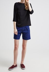 Osoba oblečená v černém topu s dlouhým rukávem, modrých šortkách po kolena a vínových nazouvacích botách, stojící na jednocenném šedém pozadí.