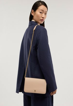 Kvinde i navy blazer med beige crossbody taske med guldkæde rem, der kigger over skulderen mod en ensfarvet baggrund.