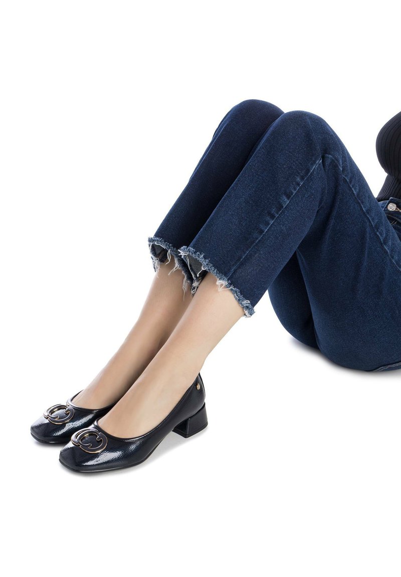 Zapatos de tacón bloque de charol negro con punta redonda y detalle de hebilla circular dorada. Llevados con jeans de denim de dobladillo deshilachado.