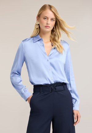Frau mit langen blonden Haaren, die eine hellblaue Seidenbluse und dunkelblaue, hoch taillierte Hose mit Gürtel trägt, posiert mit einer Hand in der Tasche.