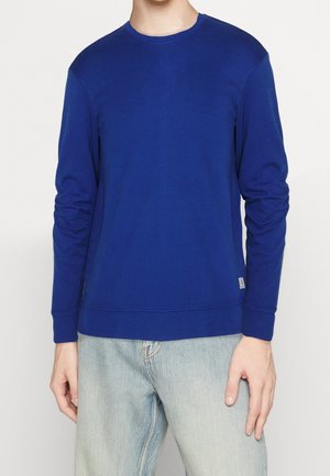 Osoba ubrana w prostą, niebieską bluzkę z długim rękawem i dekoltem typu crew oraz jasne, wypłowiałe dżinsy, stojąca na tle białego tła.