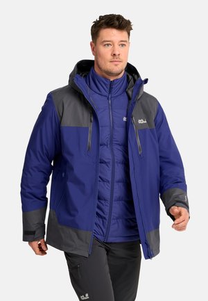 Man met kort haar die een gelaagde blauw-grijze outdoor jas en zwarte broek draagt en vooruit loopt tegen een effen achtergrond.
