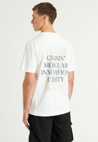 Balta medvilninė marškinėliai su grafika ant nugaros, kurioje užrašas "CHASIN' MODULAR INNOVATION UTILITY" tamsiai mėlyna spalva. Trumpomis rankovėmis.