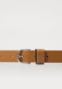 Ceinture en cuir tan avec une boucle argentée et un logo gravé. Texture lisse avec plusieurs trous pour réglage.
