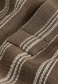 Gros plan sur un tissu marron texturé avec des coutures blanches rayées et un détail de boucle en tissu.