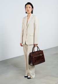 Costume beige ajusté avec un pantalon à jambes larges, associé à un sac à main en crocodile brillant marron. Des chaussures noir et blanc complètent le look.