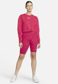 Rožinis džemperis su apvalia iškirpte ir raišteliu apačioje, derinamas su rožiniais dviračių šortais su šviesiu raštu ir mažais logotipais. Balti sportbačiai.