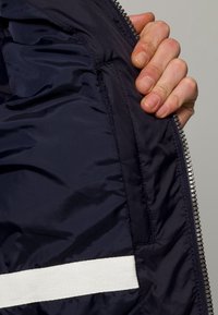 Innersidan av en marinblå isolerad jacka med slätt, glänsande tyg och vita horisontella randdetaljer. Dragkedja synlig vid kanten.