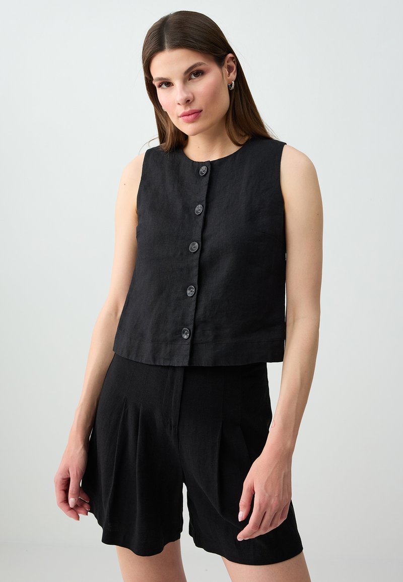 Femme aux cheveux bruns lisses portant un haut noir sans manches boutonné et un short noir assorti, debout devant un fond clair uni.