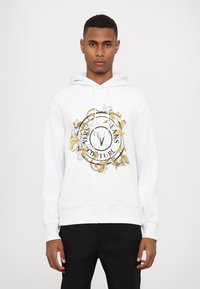 Balta hoodie su centriniu auksiniu ir juodu grafiniu motyvu, puošniais raštais ir užrašu "Versace Jeans Couture". Reguliuojama kapisono virvutė.