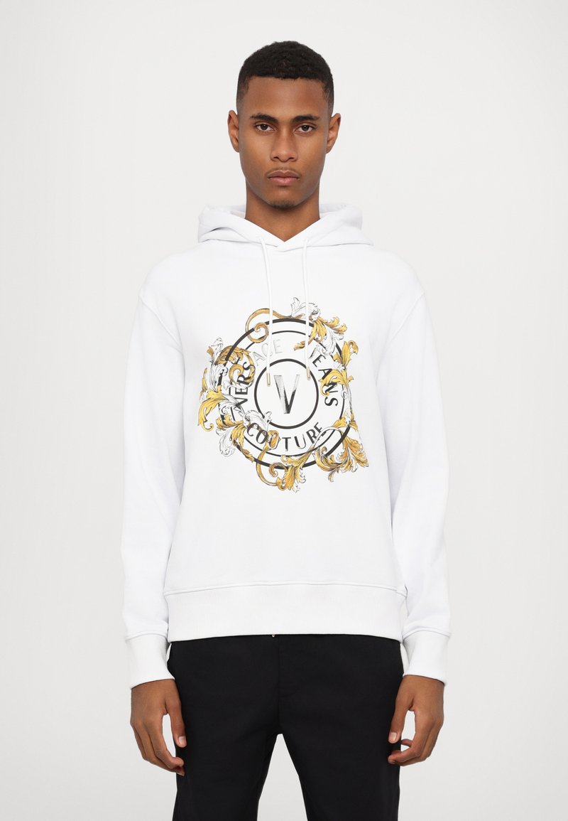Balta hoodie su centriniu auksiniu ir juodu grafiniu motyvu, puošniais raštais ir užrašu "Versace Jeans Couture". Reguliuojama kapisono virvutė.