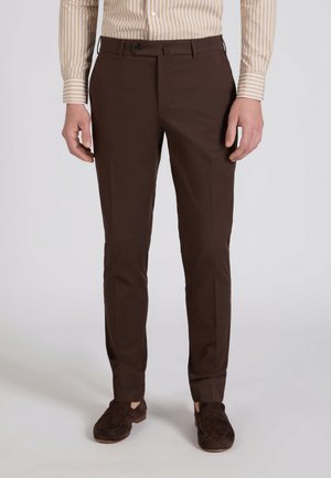 Mężczyzna ubrany w dopasowane ciemnobrązowe spodnie, beżową koszulę w paski na guziki oraz ciemnobrązowe mokasyny, stojący na prostym tle.