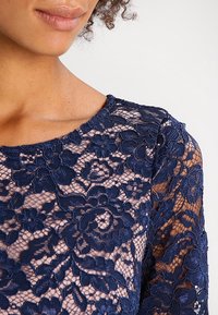 Marinblus med spets, blommönster, genomskinligt tyg, rund halsringning och långa ärmar. Texturdetaljer genom hela plagget.