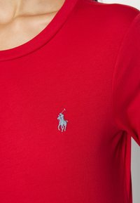 Detailní záběr na červenou košili s malým vyšitým logem polo hráče na levé hrudi.