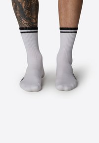 Calcetines blancos con rayas negras en el puño, hechos de un tejido elástico, que presentan lados texturizados y un logo claro en el tobillo exterior.