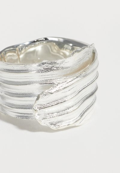 Ευρύ ασημένιο δαχτυλίδι με υφασμένη, πολυεπίπεδη επιφάνεια και κυματιστή, διπλωμένη σχεδιαστική λεπτομέρεια στη μία πλευρά.