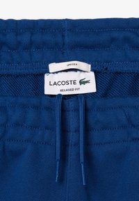 Blauwe comfortabele broek met een tailleband met trekkoord, gemaakt van zachte stof. Bevat een label met "LACOSTE" branding en een geborduurd logo.