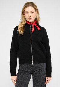 Pull noir à fermeture éclair avec un tissu texturé, des poignets et un ourlet côtelés. Associé à un jean gris à imprimé léopard et une écharpe rouge à motifs.