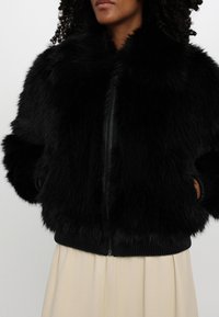 Giacca bomber in pelle sintetica nera con maniche lunghe, chiusura con zip, polsini e orlo a coste. Tessuto morbido, design voluminoso e finitura soffice.