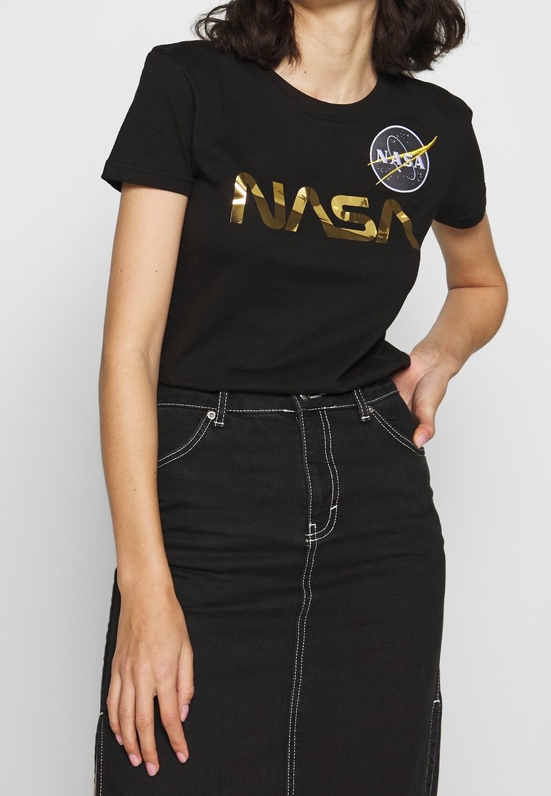 Camiseta de algodón negra con un logotipo metálico dorado de la NASA y un parche circular de la NASA. Combinada con una falda de denim negra con costuras blancas.