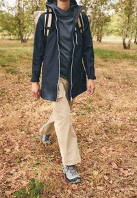 Chaqueta impermeable azul marino con cremalleras plateadas, pantalones beige y zapatos de senderismo grises. Caminando sobre hierba seca con hojas esparcidas en una zona boscosa.