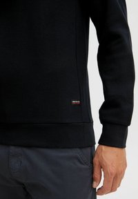 Black textured pullover with a snug fit, featuring a ribbed hem and cuffs. Small label with brand name on the lower side.