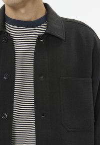Sort button-up jakke med en brystlomme, båret over en navy og hvid stribet t-shirt, som fremhæver kontrasterende teksturer og farver.