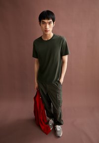 Olivegrünes T-Shirt mit kurzen Ärmeln und rundem Ausschnitt, kombiniert mit dunklen Cargo-Hosen. Hält einen roten Sweatshirt und trägt graue Turnschuhe.