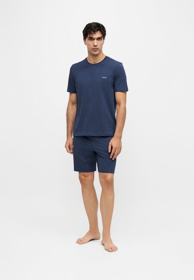 Jeune homme debout pieds nus portant une chemise bleu marine à manches courtes et un short assorti avec un petit logo "BOSS" sur la poitrine de la chemise.