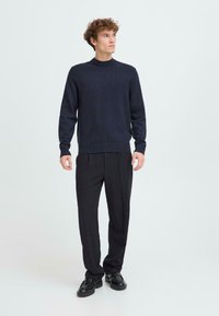 Maglione blu navy a maglia con collo a giro, abbinato a pantaloni neri plissettati e scarpe nere, che mettono in mostra una silhouette slanciata.