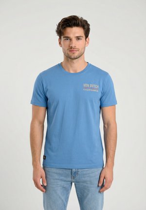 Joven de pie, mirando hacia adelante, vestido con una camiseta azul Von Dutch California y jeans azul claro, sobre un fondo liso.