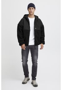 Giacca in pile nera con tessuto nero a contrasto, chiusura con zip, T-shirt bianca, jeans grigi e scarpe da trekking grigie con suole testurizzate.