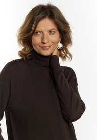 Brauner Rollkragenpullover mit lockerer Passform, strukturiertem Strick und fallenden Schultern, kombiniert mit auffälligen, runden Ohrringen.