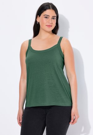 Woman with long dark hair wearing silver hoop earrings, green sleeveless top, and black pants, standing against a white background.