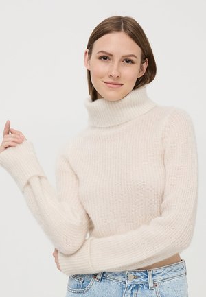 Maglione a collo alto beige chiaro realizzato in morbido materiale lavorato a maglia; presenta una texture a coste e maniche allungate, abbinato a jeans a vita alta blu chiaro.