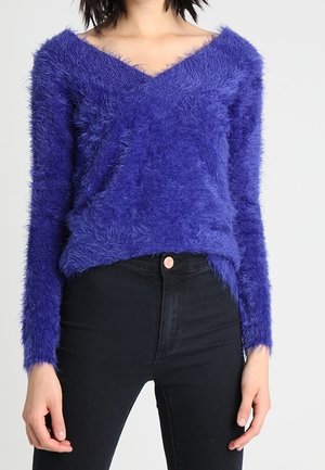 Vrouw draagt een pluizige, paarse V-hals trui met lange mouwen, ingestopt in een zwarte high-waist jeans tegen een effen achtergrond.