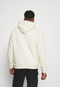 Felpa color crema con un ampio cappuccio, maniche lunghe e orlo a coste. Il materiale appare morbido con una superficie liscia, indossata con pantaloni neri.