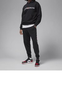 Persona che indossa un hoodie nero Jordan e pantaloni della tuta con sneakers Nike Air Jordan bianche e rosse su sfondo grigio.