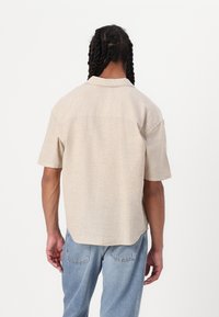 Chemise beige clair à manches courtes avec une coupe décontractée, une texture subtile et un col plat. Portée avec un jean en denim bleu clair, vue de dos.