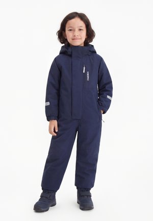 Criança com cabelo escuro vestindo um macacão de inverno azul marinho com capuz, botas e remendos refletores nas mangas.