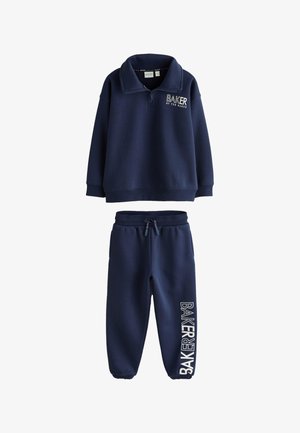 Dječak stoji okrenut leđima kameri, obučen u tamnoplavi trenirku s vertikalnim natpisom „BAKER“ na jakni i bijelim tenisicama.