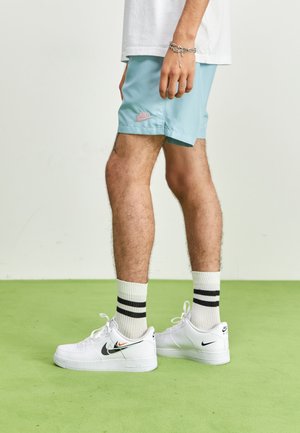 Man draagt lichtblauwe Nike-shorts, witte Nike-sneakers met zwarte swoosh, witte sokken met zwarte strepen, staat op een groene vloer.
